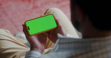 Man in casual clothes holds a mobile phone with a green screen turned horizontally while watching a indoors. Top view, no face, close-up of a man's hands photo