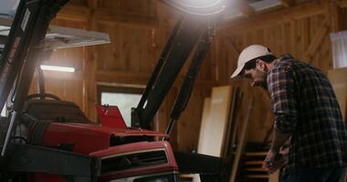 European man with a beard repairs a small agricultural machine using tools in a wooden barn, under artificial lighting photo