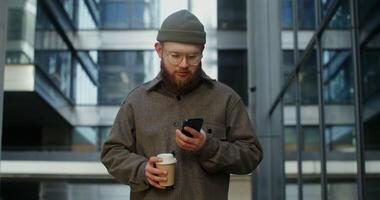 Warmly dressed bearded man in glasses is typing text on a mobile phone while standing near a stylish office building with a disposable cup in his hands. in 4k, red komodo photo