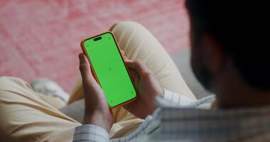 A man holds a mobile phone with green screen in his hand, sitting on the couch at home. Top view, no face, close-up of a man's hands photo
