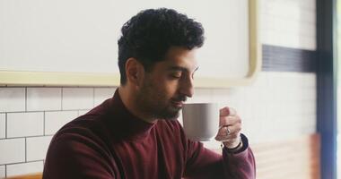 Young man with thick stubble on his face, dressed in casual clothes, enjoying the aroma of coffee inhaling deeply and taking a sip while sitting at a table in a cafe photo