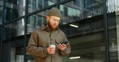 Bearded man in glasses uses a mobile phone while standing near a stylish office building with a disposable cup in his hands. Dynamic circular motion . in 4k, red komodo photo