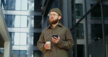 Bearded stylishly dressed man smiling while using a mobile phone while standing near an office building with large darkened windows. in 4k, red komodo photo