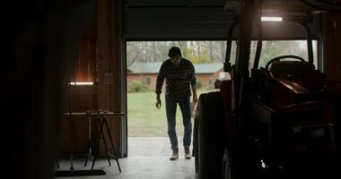 Garage door slowly opens and a young man of European appearance comes inside, inspecting the tractor parked there and getting behind the wheel. Filming from the darkness of the garage photo