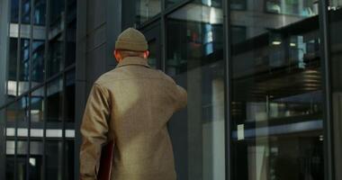 Warmly dressed man drinks hot drink from a disposable cup while walking near city buildings with a laptop in his hand, from the back of a man, his face is not visible. in 4k, red komodo photo