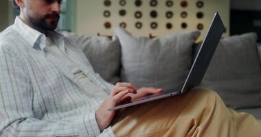 Stylish man in casual clothes starts a call on a laptop smiling and waving hello while sitting on a sofa at home photo