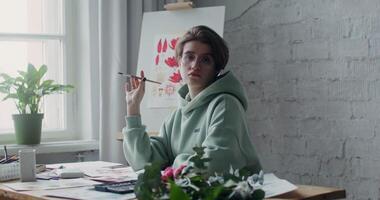 Young artist in glasses looking at the camera, while posing with a drawing brush in her hands while sitting at a desk with drawings laid out on it photo