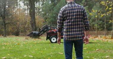 Black-haired man with a beard, dressed in casual clothes with a baseball cap on his head, walks up to the tractor and gets into it, getting ready to work in the field photo