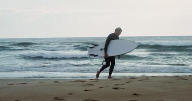 Man in a diving suit with a surfboard in his hand walks along the shore at dusk. Foam waves hit the sandy shore photo