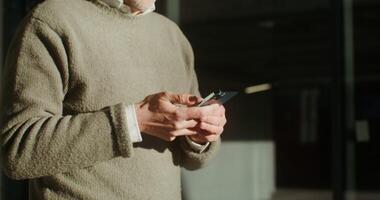 Gray-haired man dressed in sweater is typing a message on his phone during walking around the city. Close-up, the moves from the man's hands to his face. in 4k, red komodo photo