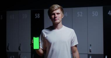 Young man, dressed in a sports uniform, smiling while looking at the camera and holding a phone with a green screen in his hands while standing in a stylish locker room. Copy space, chroma key photo