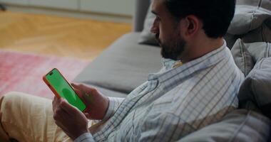 A bearded man uses a mobile phone with a green screen, scrolling the screen with his finger while sitting on a sofa indoors photo