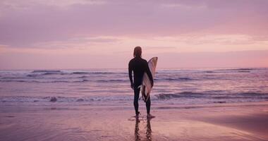 View of a man in a diving suit with wet hair and a surfboard in his hands, standing on the seashore in the pink light of the setting sun and looking into the distance photo