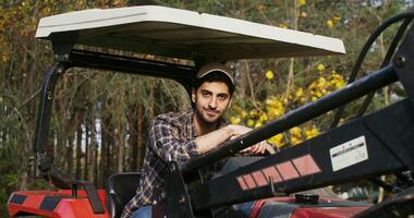 Man of Caucasian ethnicity in a baseball cap, sits with his hands on the steering wheel of an agricultural car and smiling while looking at the camera on a sunny day photo