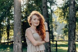 Curly redhead woman standing still in forest path with sunlight beams breaking through trees and rainbow reflection on her dress soft facial expression and peaceful presence in emotional natural photo