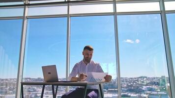 Businessman at workplace on office windows background. Handsome man in white shirt sitting at the table with a laptop and working on new project. video