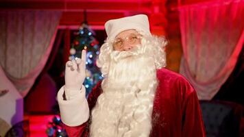 Santa Claus smiling on camera indoors. Portrait of a real Santa with white beard, in glasses and red costume standing in the living room on Christmas tree background. video