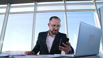Happy businessman in office on window background. Portrait of successful entrepreneur sitting at table and reading phone message. video