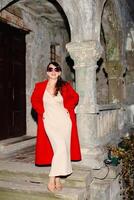 Fashion model posing in red coat and sunglasses near ancient column photo