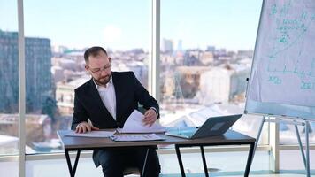 Portrait of a businessman at workplace. Bearded office worker in suit sitting at the table with a laptop on window background in the city. video