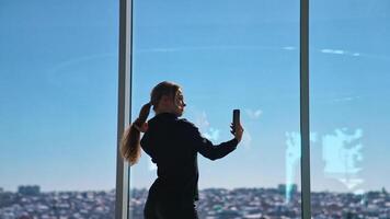 Lovely business woman in black taking photo of herself. Beautiful young woman doing selfie on smartphone while standing at office window. video