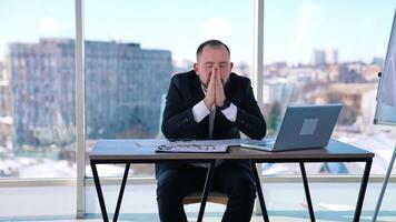 Tired businessman showing various expressions of his face. Office worker sitting at the table on window background. video