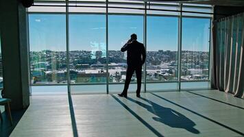 Top manager dealing with business partners through the phone. Male entrepreneur in elegant suit standing by the window and talking the phone. Panoramic windows view. video