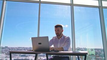 Handsome entrepreneur sitting at table in front of a laptop. Businessman working at his workplace in office on window backdrop. video