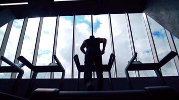 Rear view of a guy athlete exercising in gym. Dark silhouette of a sportsman running on a treadmill against sky background through windows. video