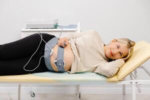 Woman pregnant checking fetal heart beat by fetal monitoring. Expectant mother with midwife adjusting sensors off cardiotocography. photo
