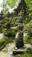 Medium shot captures a collection of weathered concrete statues depicting deities, mythical creatures, and humans in a lush, overgrown garden setting. Moss and lichen cover the sculptures video