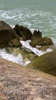 High-angle, medium shot captures dark, jagged rocks emerging from the turquoise sea along the coast of Thailand. White waves crash against the formations under an overcast sky video