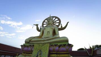 A low-angle, medium shot captures the impressive golden statue of the Big Buddha seated in a meditative pose atop a colorful lotus-shaped base. The serene Buddha figure is adorned video
