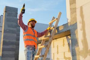 Construction worker using cordless drill on wooden ladder at building site. photo