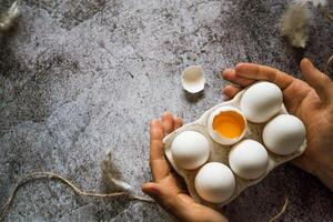 a man holds in his hands fresh farm white chicken eggs on a stand, one broken photo
