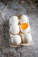 6 chicken eggs standing with feathers in a ceramic stand on a gray background, one broken egg photo
