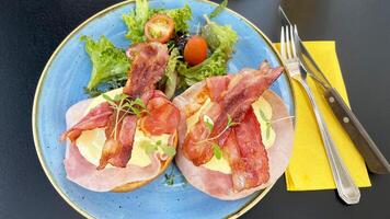 eggs benedict served with crispy bacon, ham and hollandaise sauce on brioche bun and side salad for breakfast or brunch photo