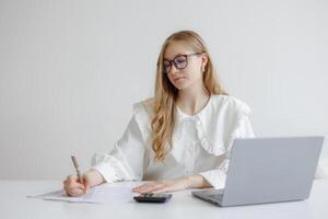 Young woman working at desk with laptop and calculator while taking notes in a bright, modern workspace during the day photo