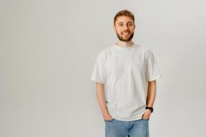 Young man stands casually in a simple white t-shirt with hands in pockets, smiling against a neutral background photo