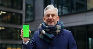 Solid warmly dressed middle-aged businessman smiling looking at camera holding the phone in his hands with the screen facing forward. Green screen, copy space, chroma key. in 4k, red komodo photo