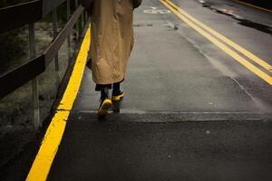 A woman in a long coat, boots walks on a wet bridge with yellow road markings. A person walks along a wet bridge, wearing a long coat and stylish boots with yellow soles. Autumnal strolling outdoors. photo