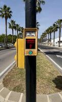 Malaga, Spain, May 18, 2025. Pedestrian traffic signal button in yellow casing instructs to press for crossing, set against a street lined with palm trees and clear blue sky, indicating urban life photo
