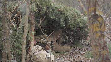 Man sheltering under pine branches during snowfall in forest, Survivalist hides from snow under handmade pine shelter with gear in cold woodland video