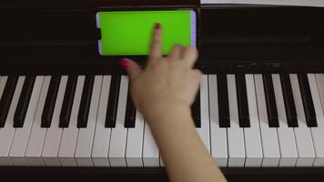A woman learns to play the piano from notes recorded in her phone. Close-up of a piano, female hands and a phone with a green screen video
