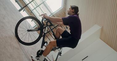 Young focused man rides a bike simulator in modern home interior. Vertical filming transforms into horizontal, circular camera movement photo