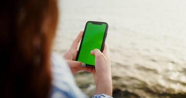 Close-up of phone with a green screen in the hands of a woman typing on a smartphone while sitting on the rocks on the seashore. Chroma key, mockup, copy space. 4k , red komodo photo