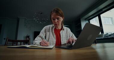 Woman makes notes in a notebook and types on a laptop, sitting at a table in a home interior with a serious look photo