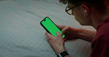 Man is typing on a smartphone with a green screen while lying on his stomach on the bed, top view. Chroma key, copy space photo
