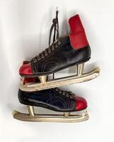 A pair of classic vintage ice skates, featuring striking black leather boots with bold red accents on the toe and heel, are artfully suspended against a clean white background. The worn appearance of photo