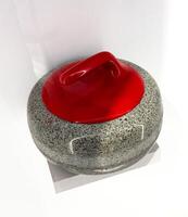 This high-angle, close-up shot captures a traditional curling stone with a vibrant red handle, set against a pristine white background. The granite body of the stone shows its unique speckled texture photo